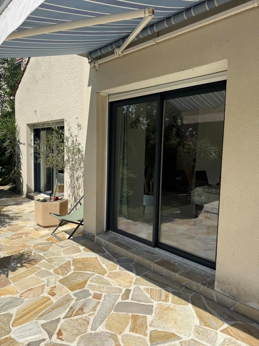 Grande baie coulissante en aluminium gris ouvrant sur une terrasse en pierre naturelle sous store